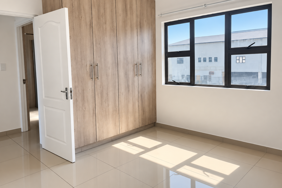A bright, empty bedroom featuring modern wood-finish built-in wardrobes, neutral tiled flooring, and large aluminium-framed windows allowing for ample natural light.