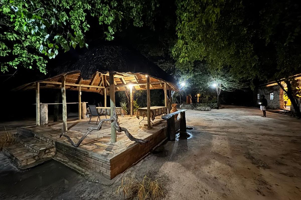 A rustic outdoor entertainment area featuring a thatched roof, stone paving, and timber supports, illuminated for evening use in a farm setting.