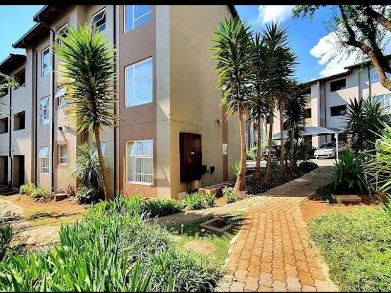 Exterior view of a multi-story apartment complex featuring a paved walkway, landscaped garden areas, and covered parking bays.