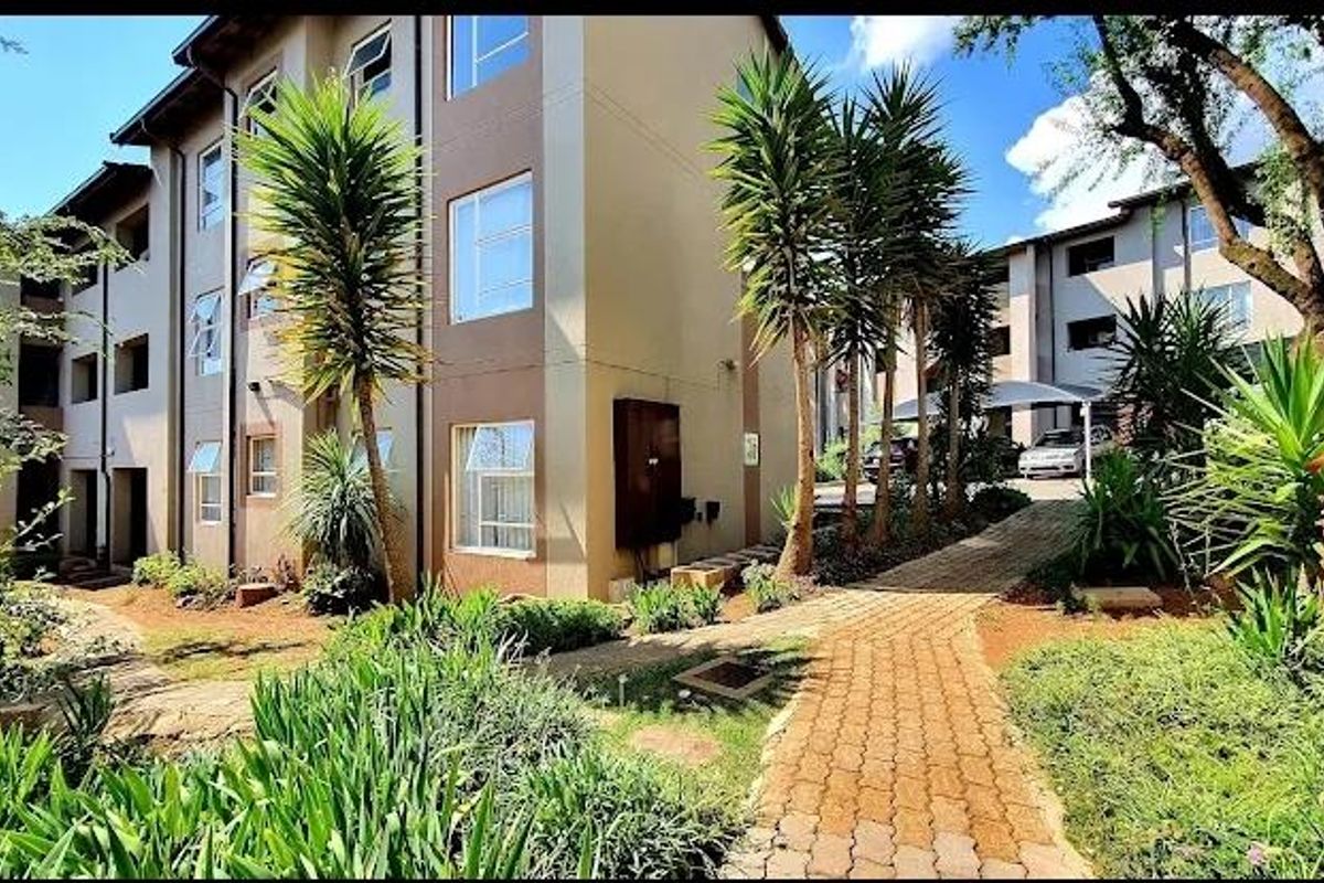 Exterior view of a multi-story apartment complex featuring a paved walkway, landscaped garden areas, and covered parking bays.
