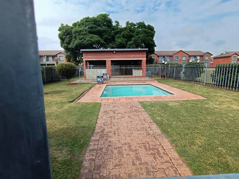 Communal swimming pool area featuring a brick-paved walkway, landscaped lawn, and a covered brick lapa structure within a secure fenced complex.