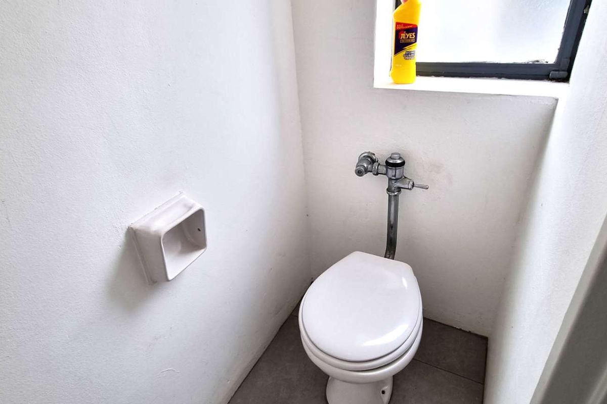 A compact industrial-style restroom featuring a wall-mounted flush valve toilet and tiled flooring.