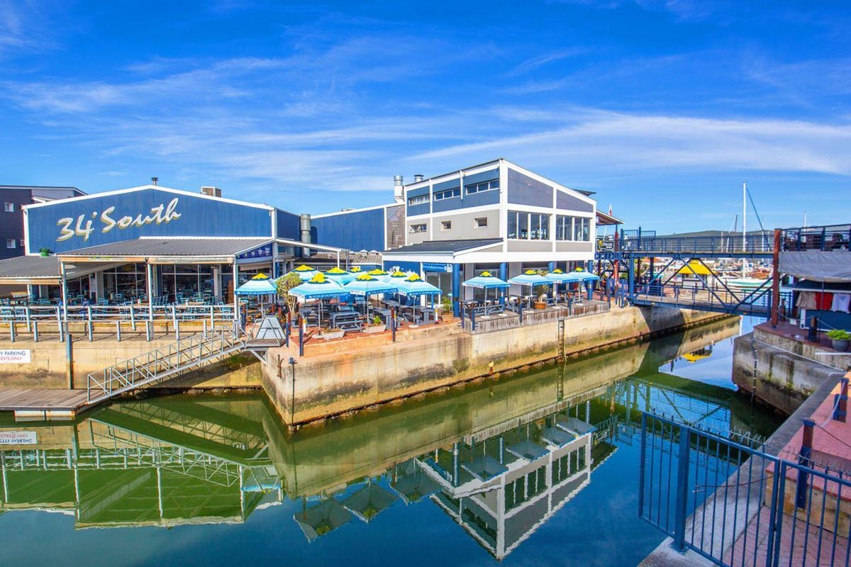 Exterior view of a commercial waterfront building featuring a concrete quay, metal railings, and a pedestrian bridge overlooking the marina.