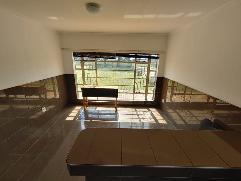 An empty living area featuring dated brown wall and floor tiles, a built-in counter, and a large window overlooking the garden.