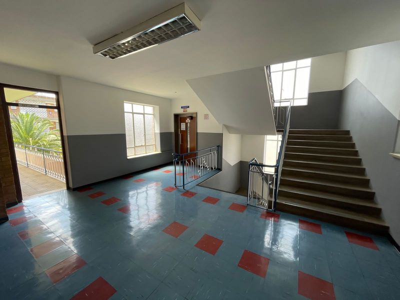 A spacious apartment building hallway featuring a staircase, elevator access, and durable tiled flooring with natural light from large windows.