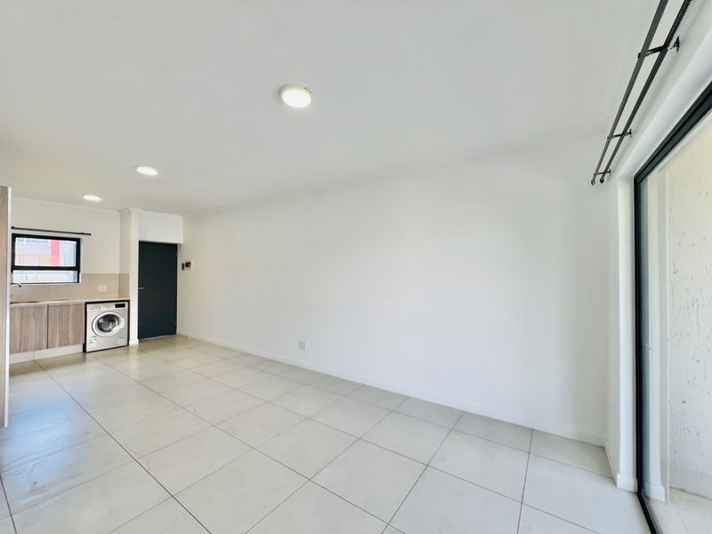 A bright, open-plan living area featuring neutral tiled flooring, recessed lighting, and a compact kitchen corner with built-in cabinetry.