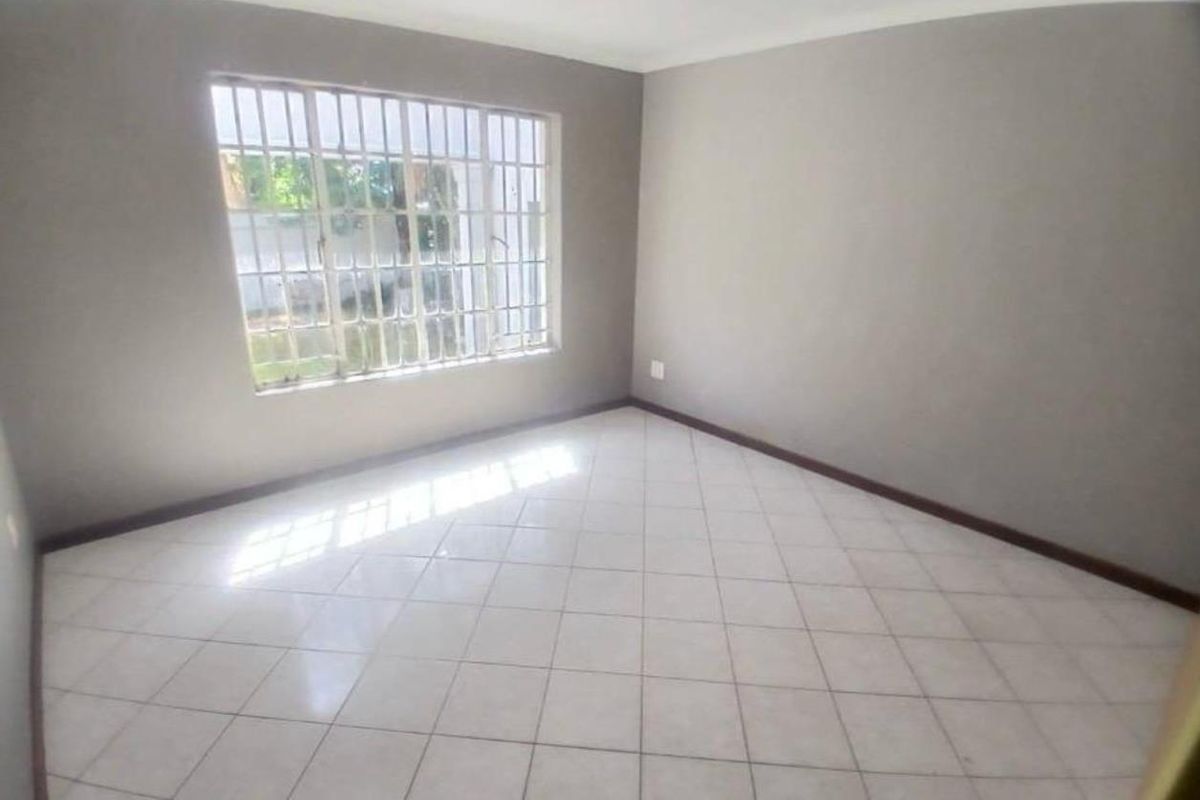 A bright, empty bedroom featuring neutral grey walls, tiled flooring, and a large window fitted with security bars.