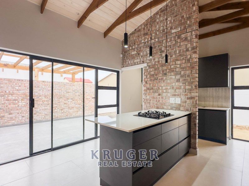 Modern open-plan kitchen and living area featuring exposed timber rafters, a striking brick feature wall, sleek cabinetry, and large sliding doors leading to the patio.