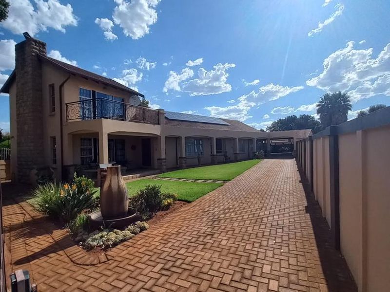 A well-maintained residential property featuring a long paved driveway, stone chimney, upper-level balcony, and solar panels under a clear blue sky.