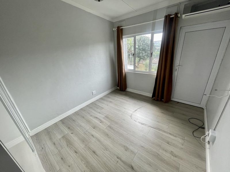 A bright, empty bedroom featuring light-toned laminate flooring, an installed air conditioning unit, and large windows providing natural light.