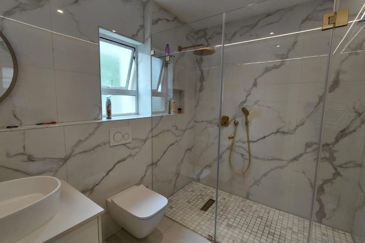Modern bathroom featuring marble-look wall tiles, a frameless glass walk-in shower with gold fixtures, a wall-mounted toilet, and a sleek vanity basin.
