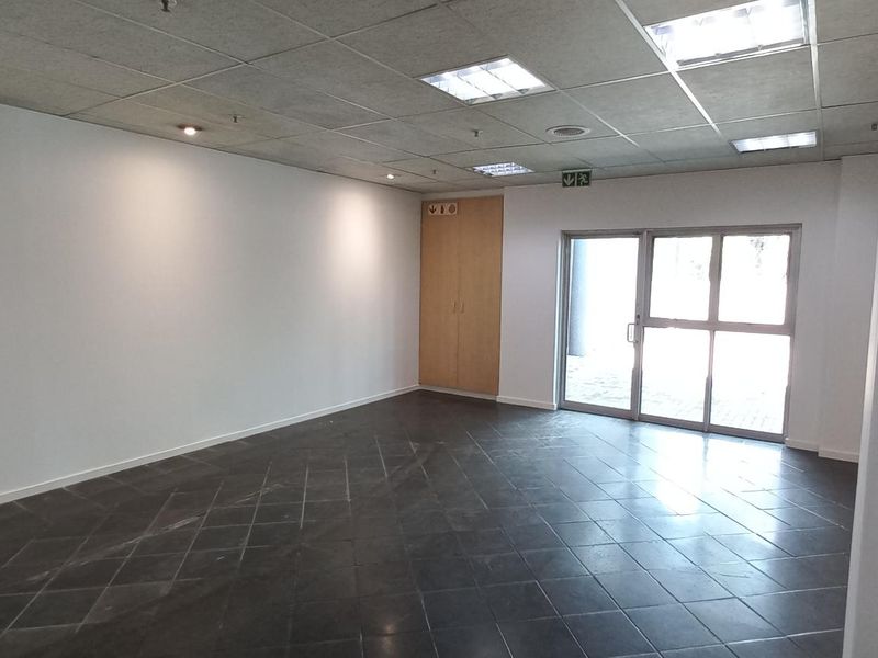An open-plan office space featuring tiled flooring, a suspended ceiling with recessed lighting, and a built-in storage cupboard with direct exterior access.