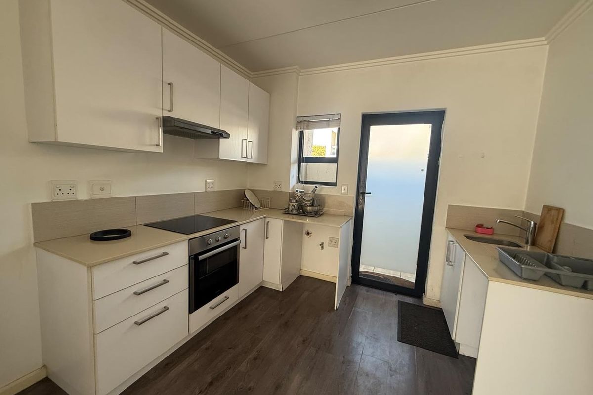 A clean, functional kitchen featuring white cabinetry, integrated oven and hob, laminate flooring, and direct access to an exterior area via a glass door.