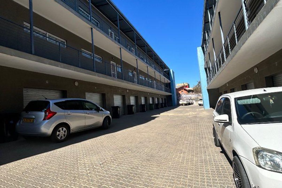 Exterior view of an apartment complex featuring a paved driveway, multiple garage units with roller shutters, and multi-level balconies with metal railings.