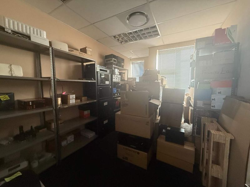 A functional utility or storage room featuring a suspended ceiling, recessed lighting, and window blinds, currently utilized for equipment and supply storage.