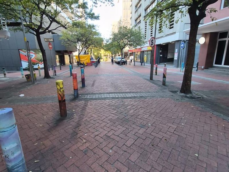 A view of a pedestrianized street area featuring brick paving, decorative bollards, and mature trees between office building facades.
