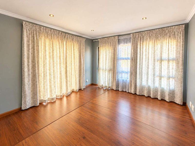 A bright and spacious living area featuring warm-toned laminate flooring, recessed downlighting, and large windows dressed with curtains.