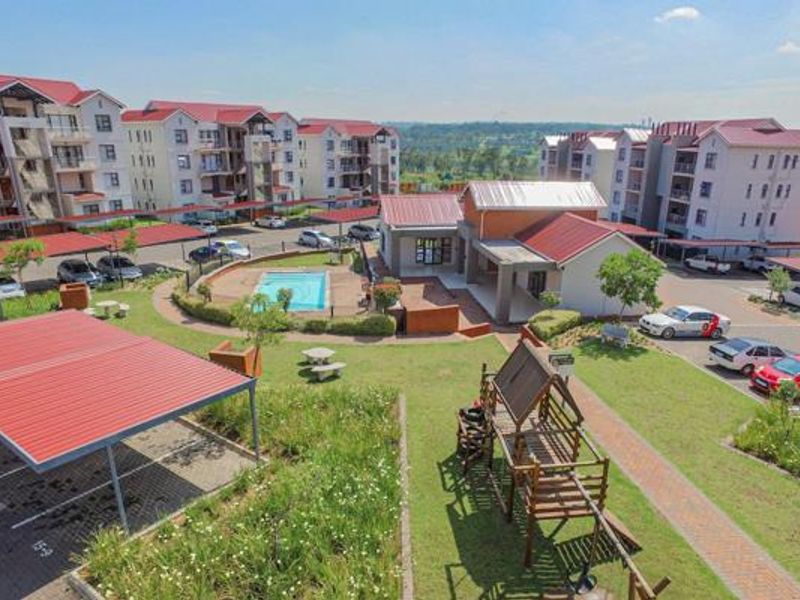 Aerial view of an apartment complex featuring communal swimming pool, landscaped gardens, paved walkways, and covered parking facilities.