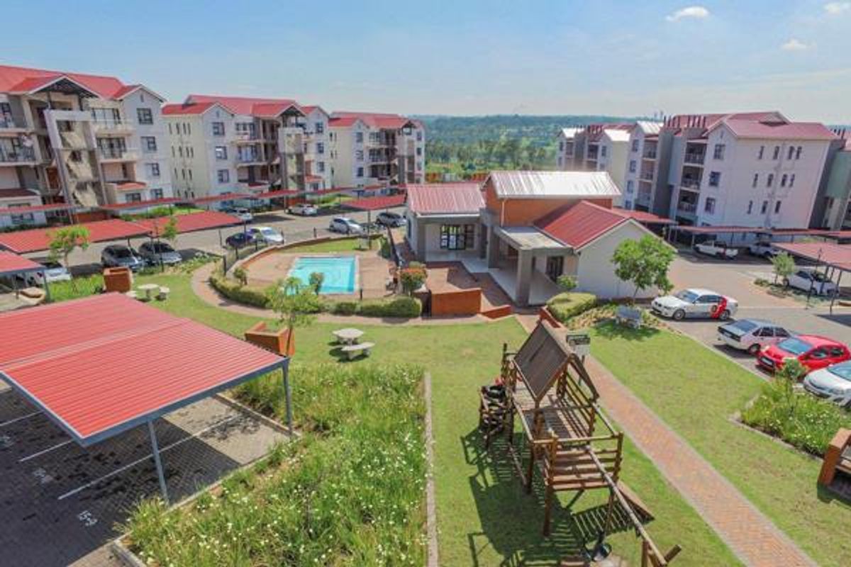 Aerial view of an apartment complex featuring communal swimming pool, landscaped gardens, paved walkways, and covered parking facilities.