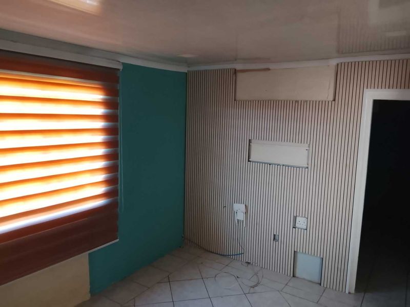 An empty living area featuring slatted wood wall paneling, tiled flooring, and modern roller blinds, currently requiring some finishing work.