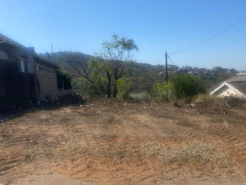 A vacant residential plot of land featuring cleared ground, adjacent to existing residential structures and local utility infrastructure.