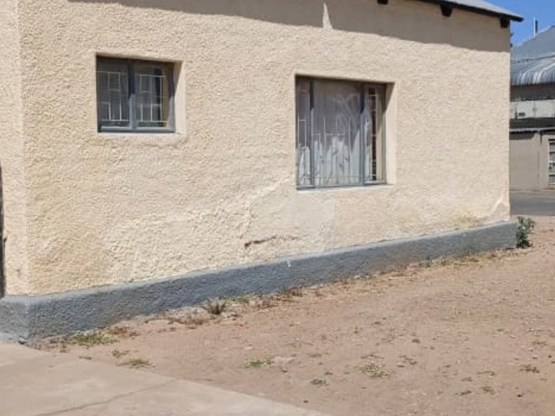 Exterior view of a building featuring plastered walls, security bars on windows, and a corrugated metal roof, showing signs of wear and weathering.