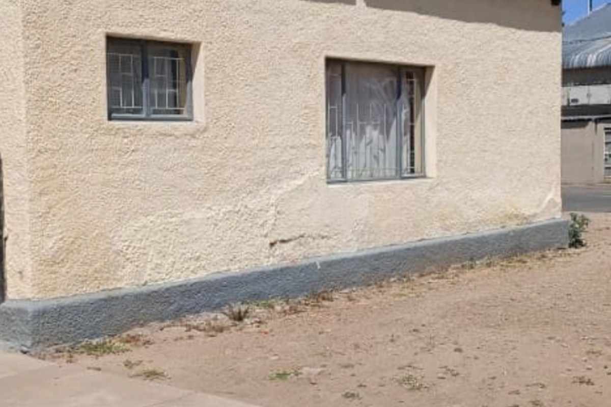 Exterior view of a building featuring plastered walls, security bars on windows, and a corrugated metal roof, showing signs of wear and weathering.