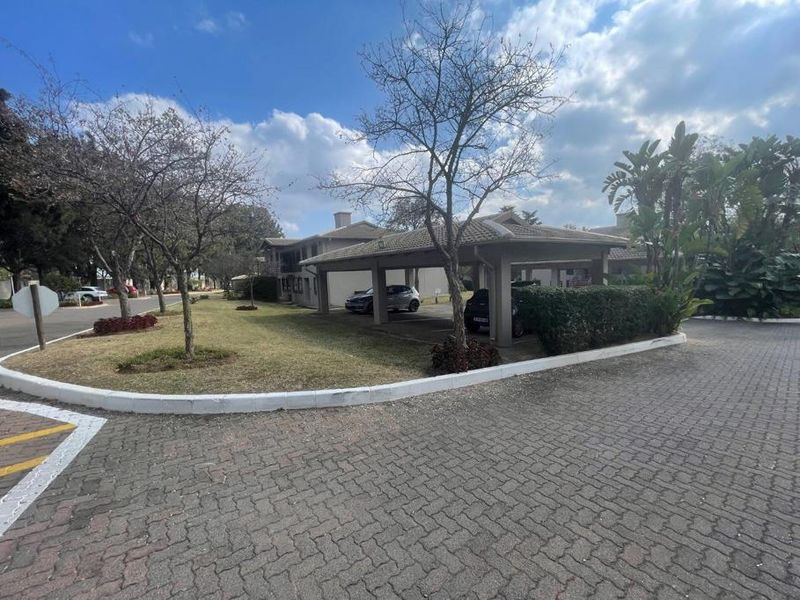 Exterior view of the apartment complex featuring a paved driveway, landscaped garden areas, and covered parking bays.
