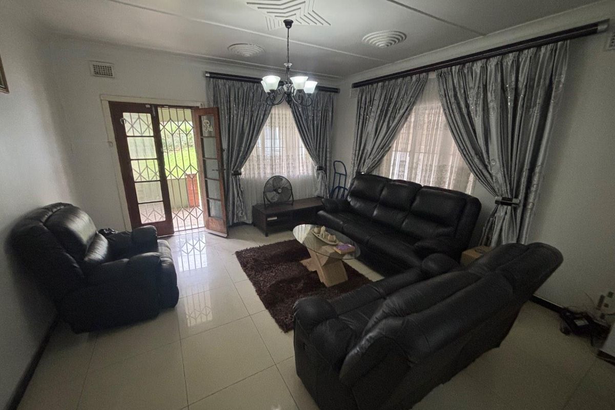 A spacious living room featuring tiled flooring, decorative ceiling molding, a central chandelier, and direct access to the exterior through a security-gated door.