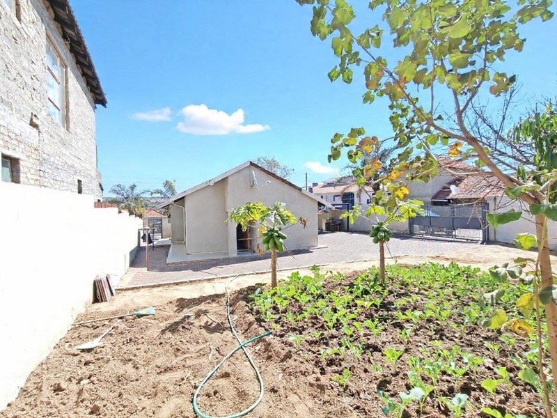 Rear view of the property featuring a paved driveway, boundary wall, metal gate, and an unlandscaped garden area with young fruit trees.