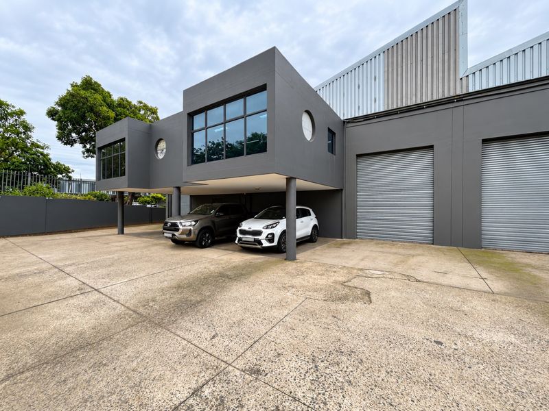 Modern mixed-use building featuring a concrete driveway, roller shutter doors, and contemporary architectural design with large glass windows and steel support columns.
