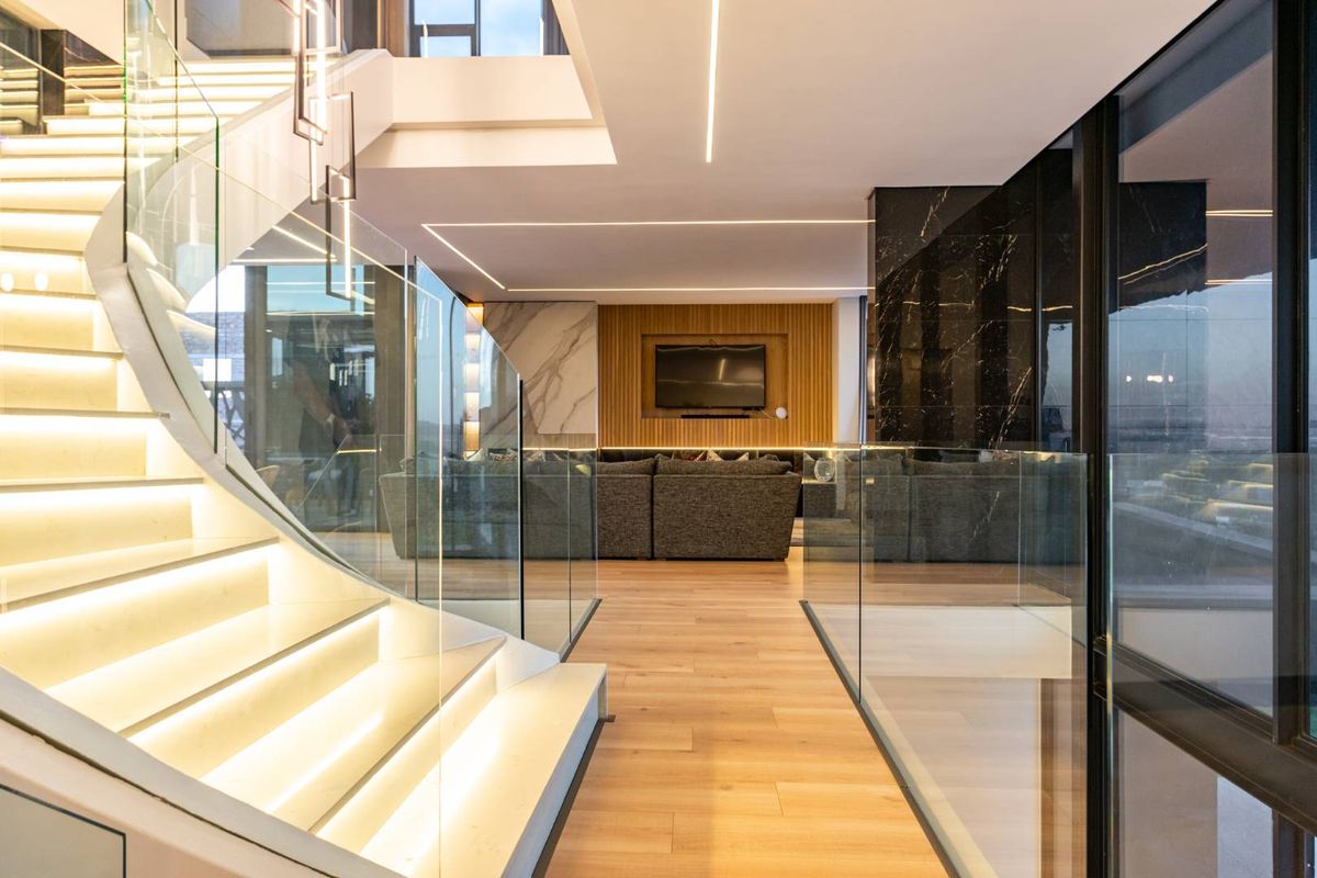 Modern open-plan living area featuring a striking illuminated floating staircase with glass balustrades, sleek timber flooring, and integrated recessed lighting.