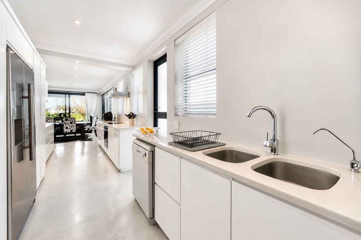 Modern galley-style kitchen featuring sleek white cabinetry, stone benchtops, stainless steel appliances, and polished concrete flooring, leading into an open-plan dining area.