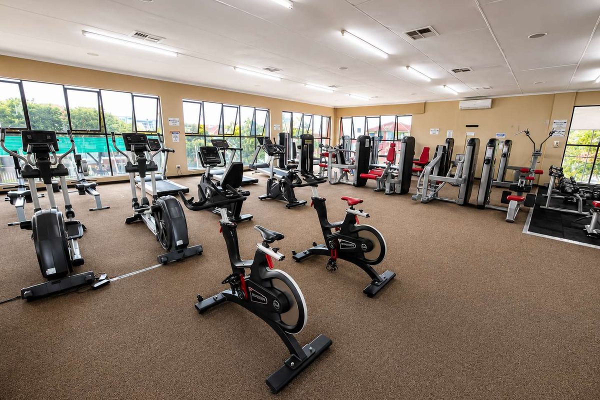 A spacious, well-lit communal gym facility featuring large windows, carpeted flooring, and a variety of fixed exercise equipment.