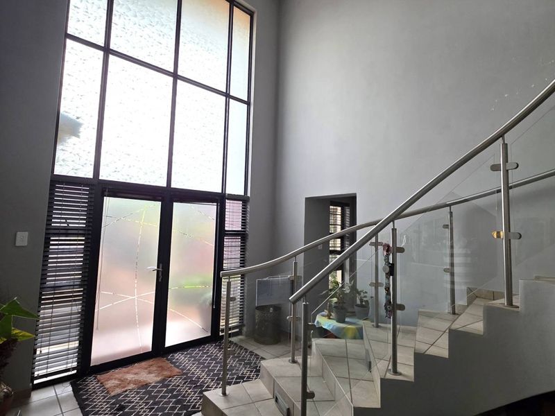 Spacious double-volume entrance hall featuring a modern glass and steel staircase, large floor-to-ceiling windows, and tiled flooring.
