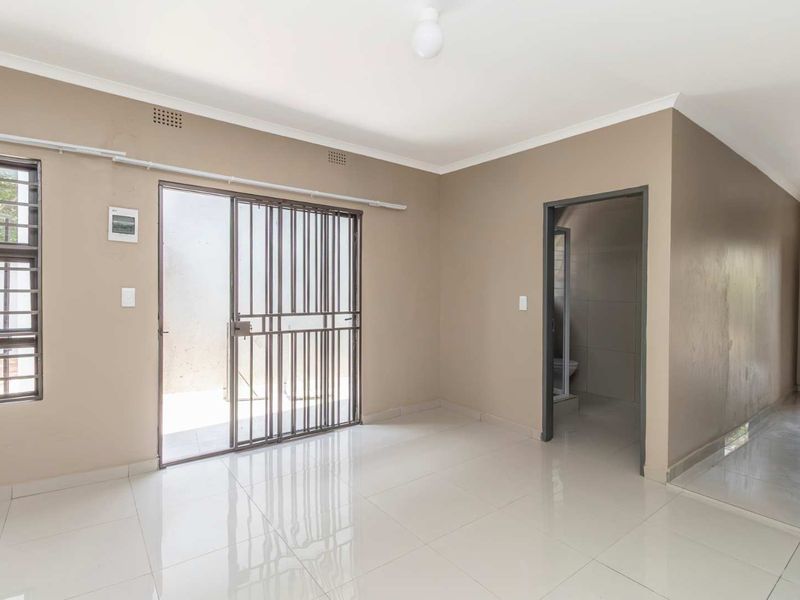 A bright, empty living area featuring neutral wall tones, polished tiled flooring, and a sliding security gate leading to the exterior.