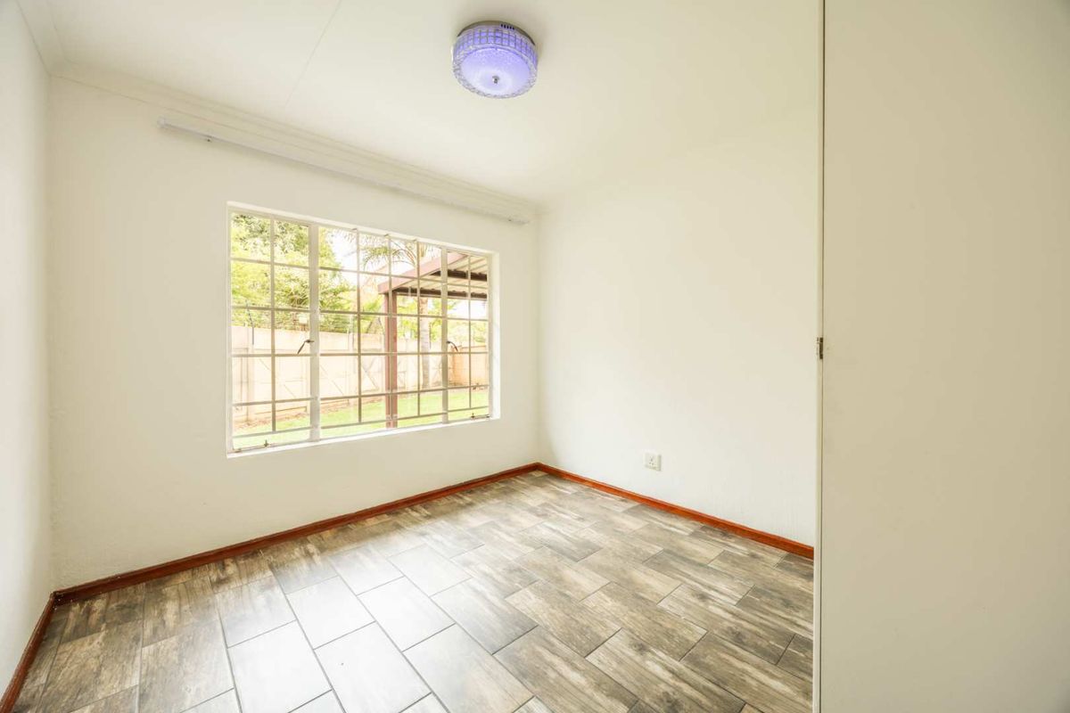 A bright, empty bedroom featuring wood-look tiled flooring, a large window with security bars, and a built-in cupboard.