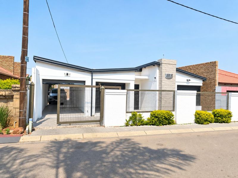 Modern single-story house facade featuring a white rendered finish, stone cladding detail, paved driveway, and secure perimeter fencing with a metal gate.