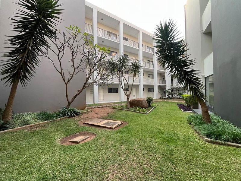 A view of the communal garden area situated between apartment blocks, featuring landscaped lawn, mature trees, and multi-level building facades with balconies.