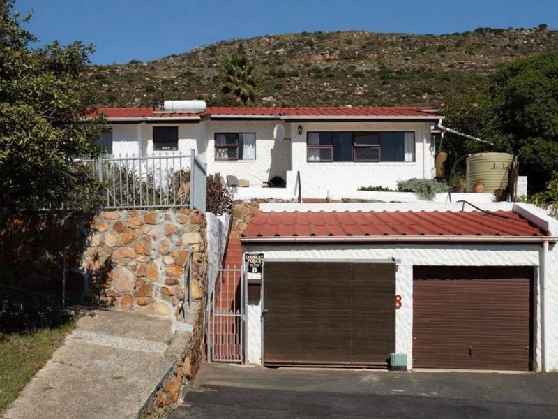 Front elevation of a residential property featuring a double garage, stone retaining wall, and a metal roof, set against a hillside backdrop.