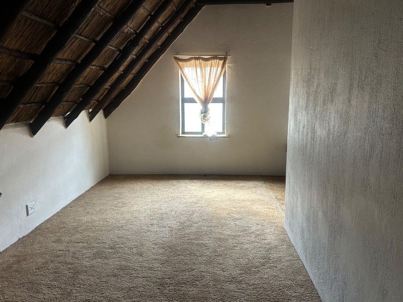An empty attic bedroom featuring a pitched thatched roof, exposed timber rafters, and carpeted flooring with a single window.
