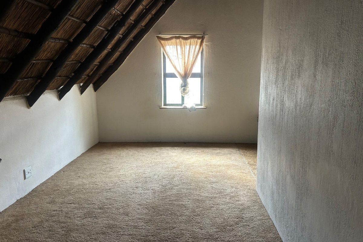 An empty attic bedroom featuring a pitched thatched roof, exposed timber rafters, and carpeted flooring with a single window.
