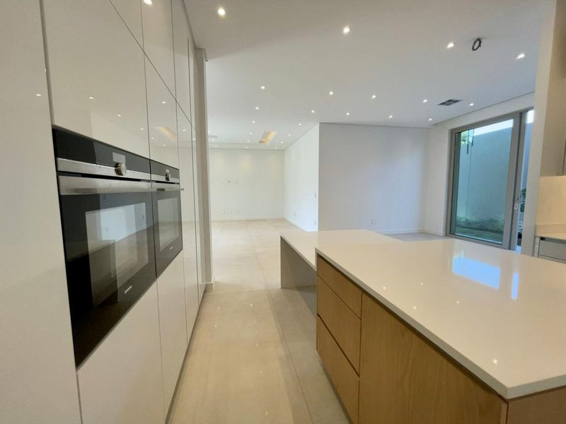 Modern open-plan kitchen featuring high-gloss cabinetry, integrated double ovens, stone countertops, and a central island, flowing into a spacious living area.