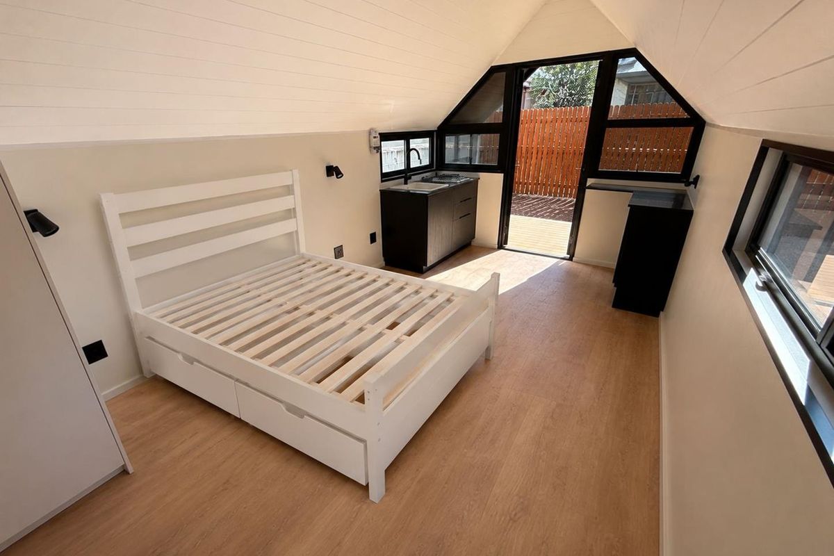 A modern, light-filled bedroom featuring a vaulted ceiling, timber-look flooring, and a compact kitchenette, with direct access to an outdoor patio area.