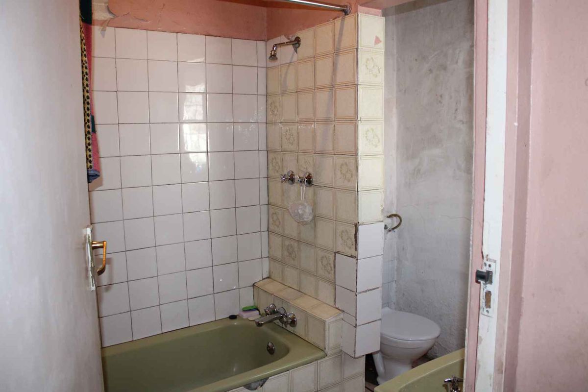 A dated bathroom featuring a bathtub with shower mixer and a separate toilet area, requiring significant renovation and cosmetic updates to improve condition.