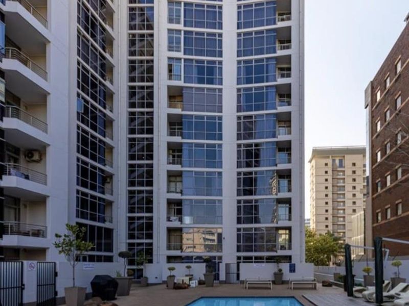 A well-maintained apartment complex pool area featuring a paved deck, timber accents, and modern glass-fronted building architecture.