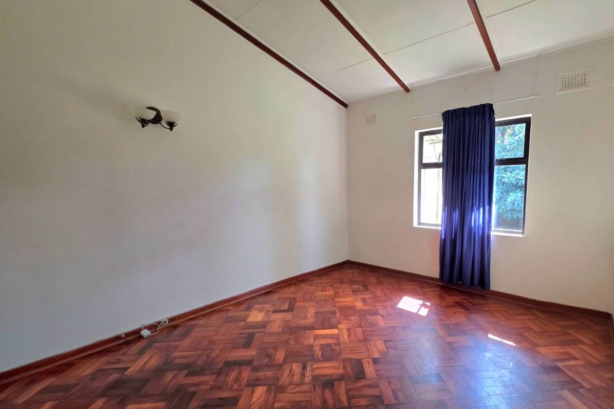 A bright, empty bedroom featuring classic parquet flooring, exposed ceiling beams, and a large window allowing for natural light.