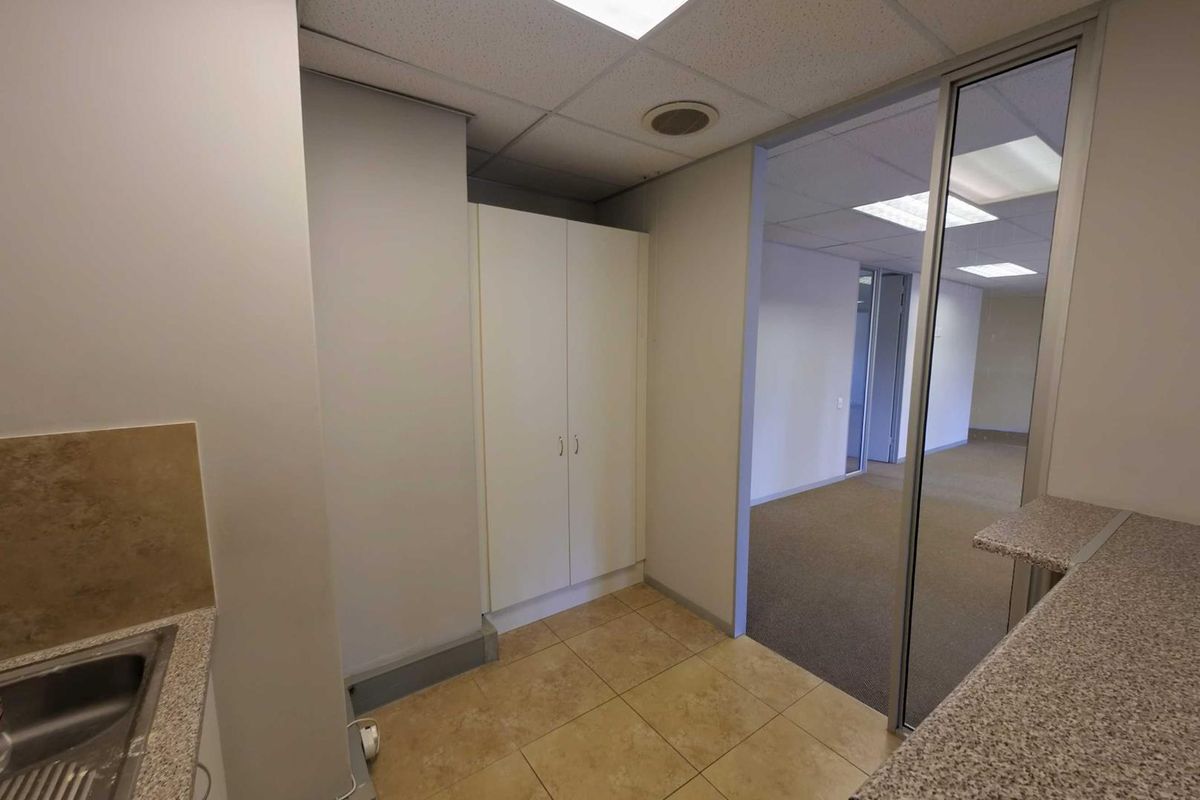A functional office kitchenette area featuring tiled flooring, built-in cabinetry, and a sink, adjacent to an open-plan office space.