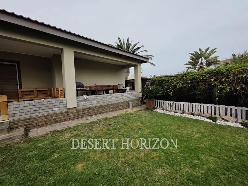 Rear view of the property featuring a covered patio with a brick retaining wall, overlooking a private garden with a lawn and timber fencing.