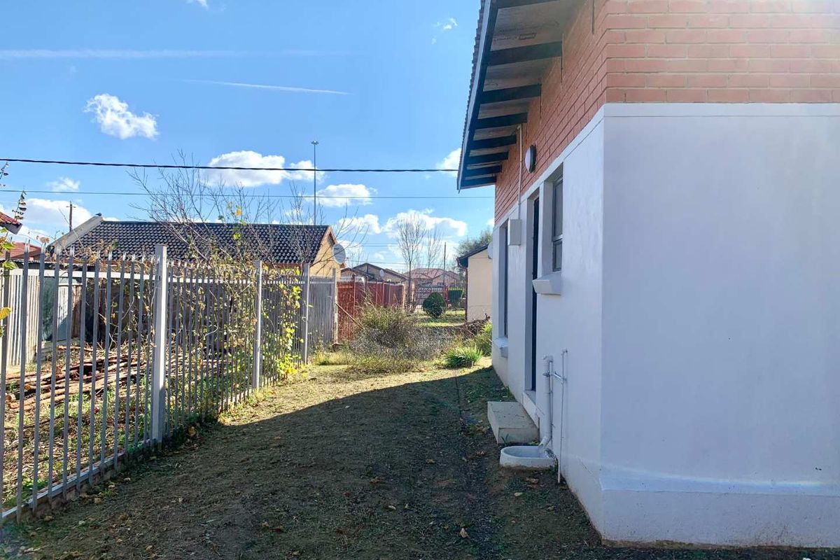 A view of the rear exterior of the house featuring a brick and painted facade, exposed roof rafters, and metal perimeter fencing.